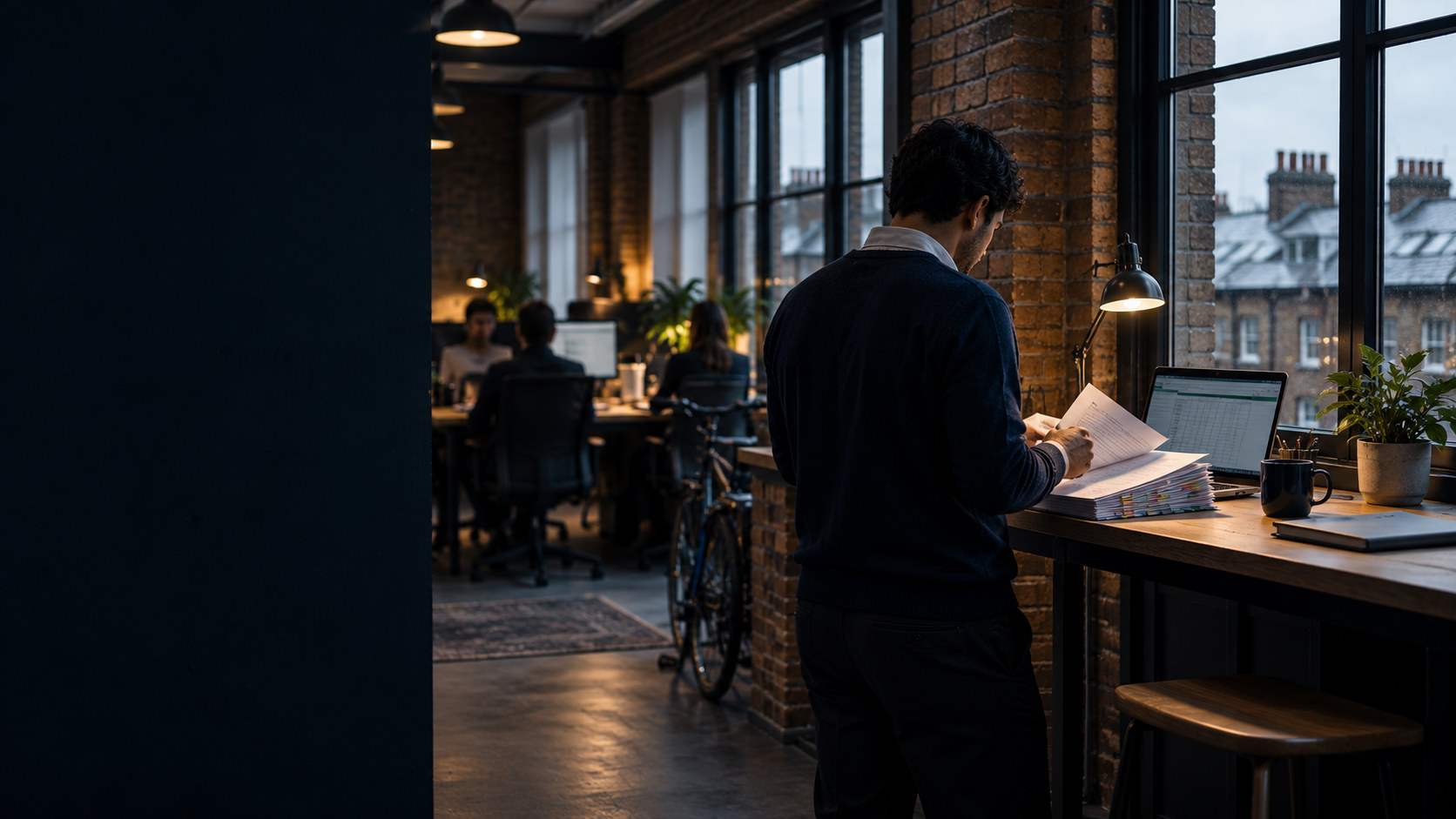 A London office at dusk: someone reading a printed finance pack at a standing desk